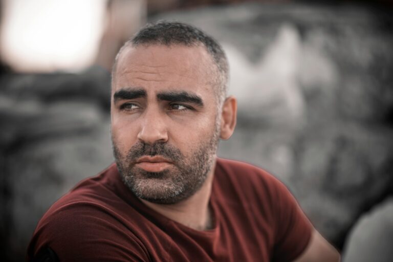 A bearded man wearing a red shirt looks to the side, set against a blurred outdoor backdrop.